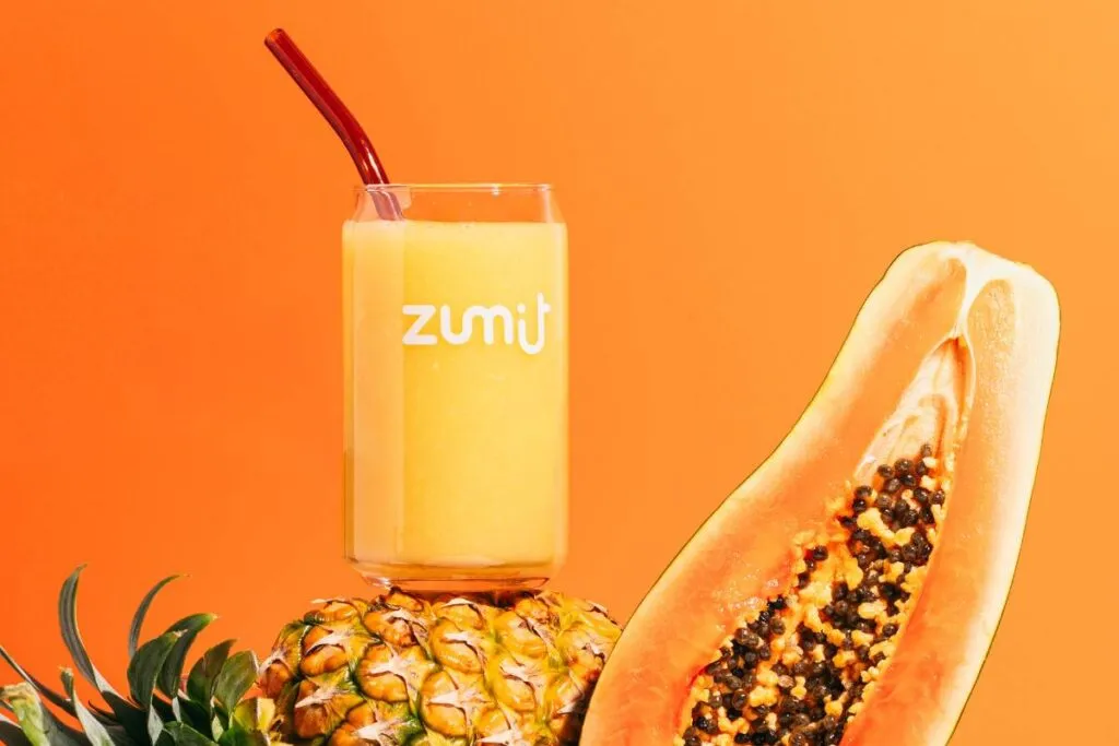 Smoothie Zumit Caribbean Passion con mango, papaya y piña apoyado sobre una piña y una papaya